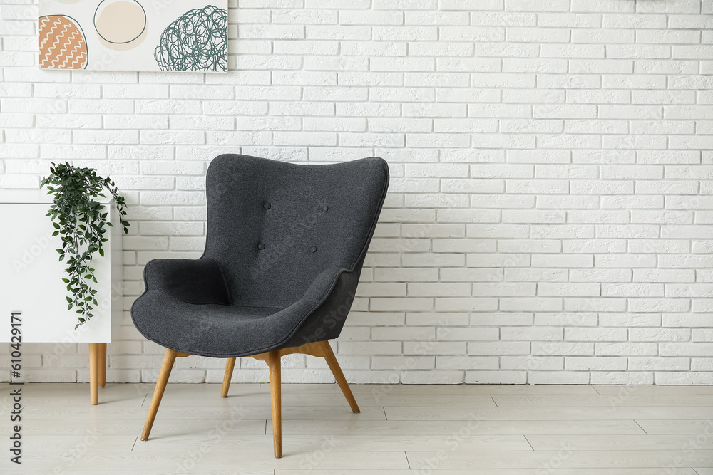 Dark grey armchair with dresser, painting and houseplant near white brick wall