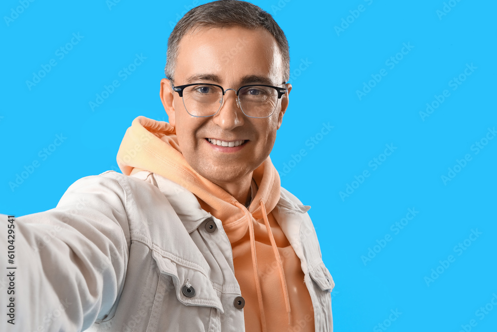 Mature man in eyeglasses taking selfie on blue background, closeup