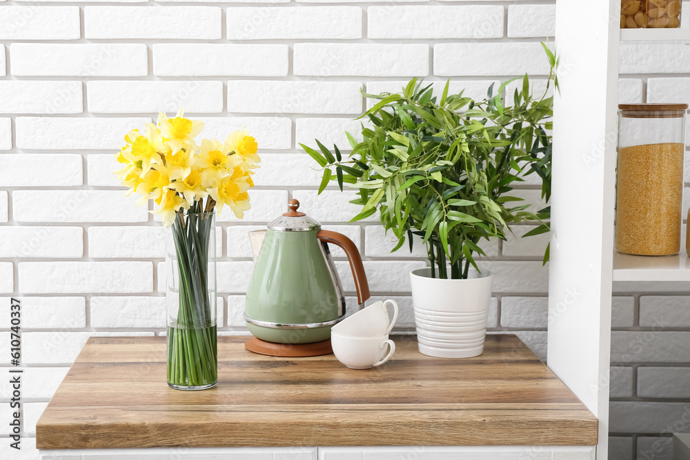 Vase with blooming narcissus flowers, electric kettle, cups and houseplant on kitchen counter