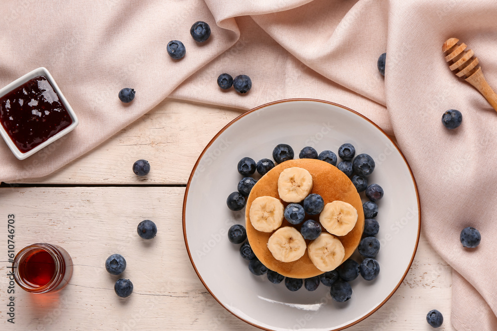 Plate with sweet pancakes, banana, blueberry and jam on light wooden background