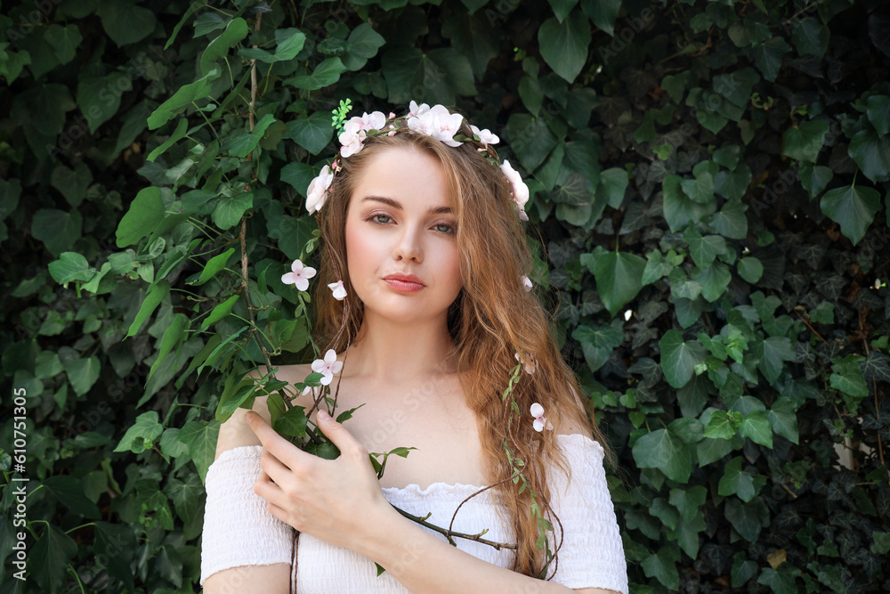 Beautiful young woman with flowers in her hair near green ivy outdoors. Summer solstice