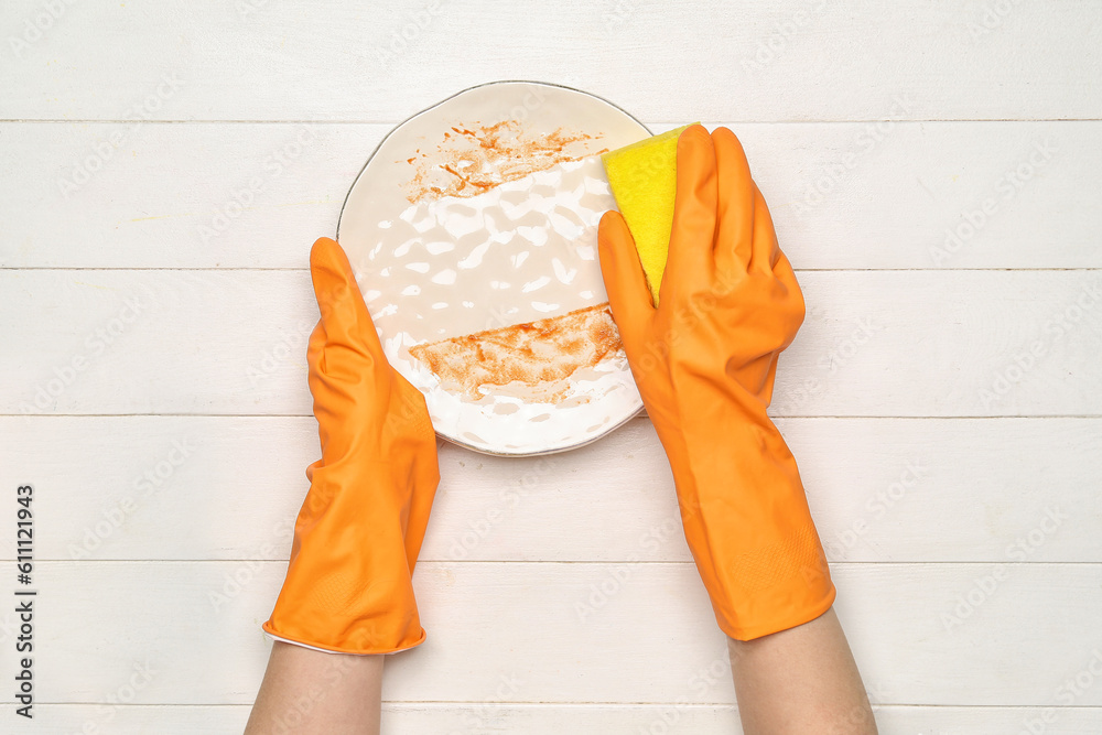 Female hands in rubber gloves washing dirty plate on white wooden background