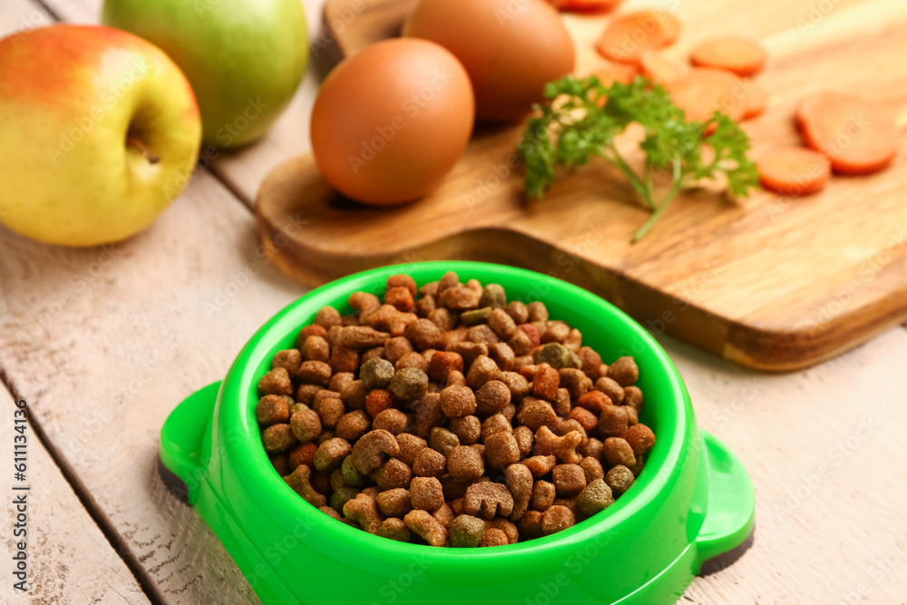 Bowl with dry pet food and natural products on light wooden background, closeup