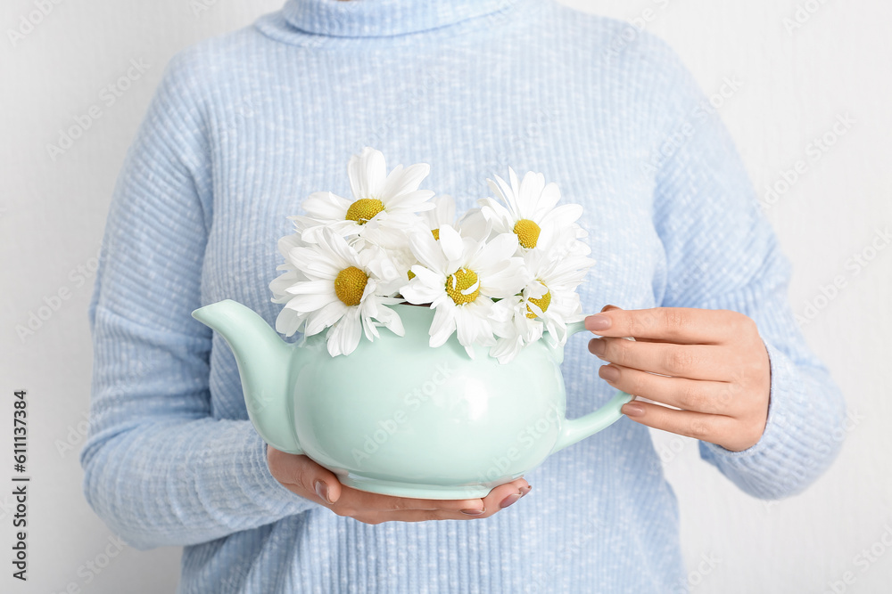 Beautiful young woman with teapot of chamomile tea and flowers near white wall