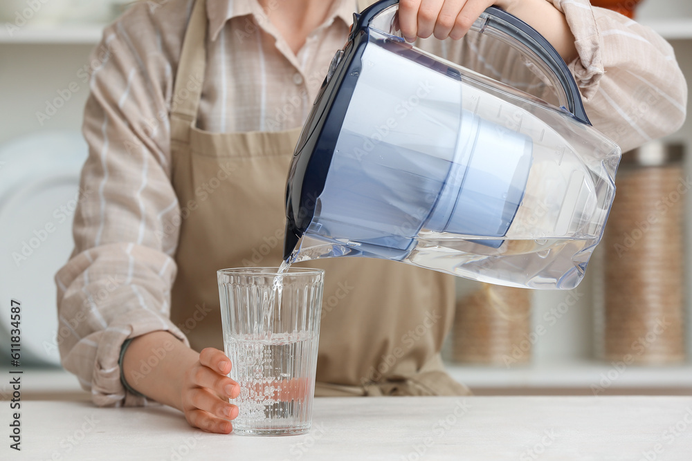 Woman pouring water into glass from filter jug at table in kitchen