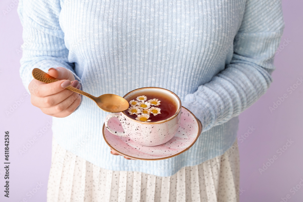 Beautiful young woman with cup of chamomile tea near lilac wall