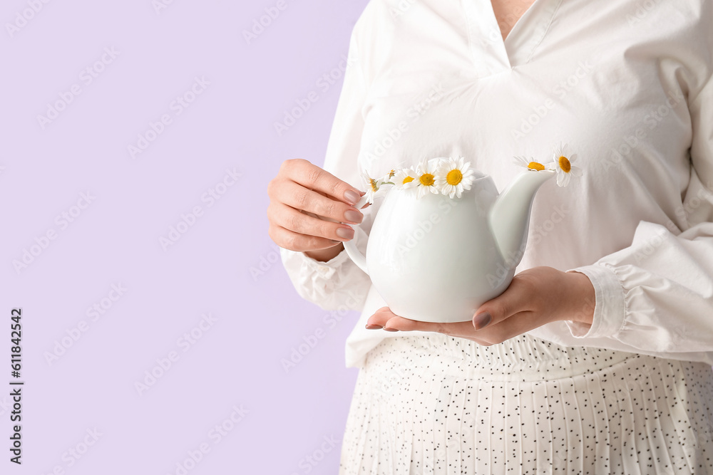 Beautiful young woman with teapot of chamomile tea and flowers near lilac wall