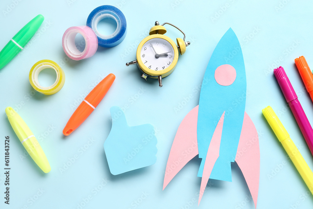 Paper rocket with alarm clock and different stationery on blue background