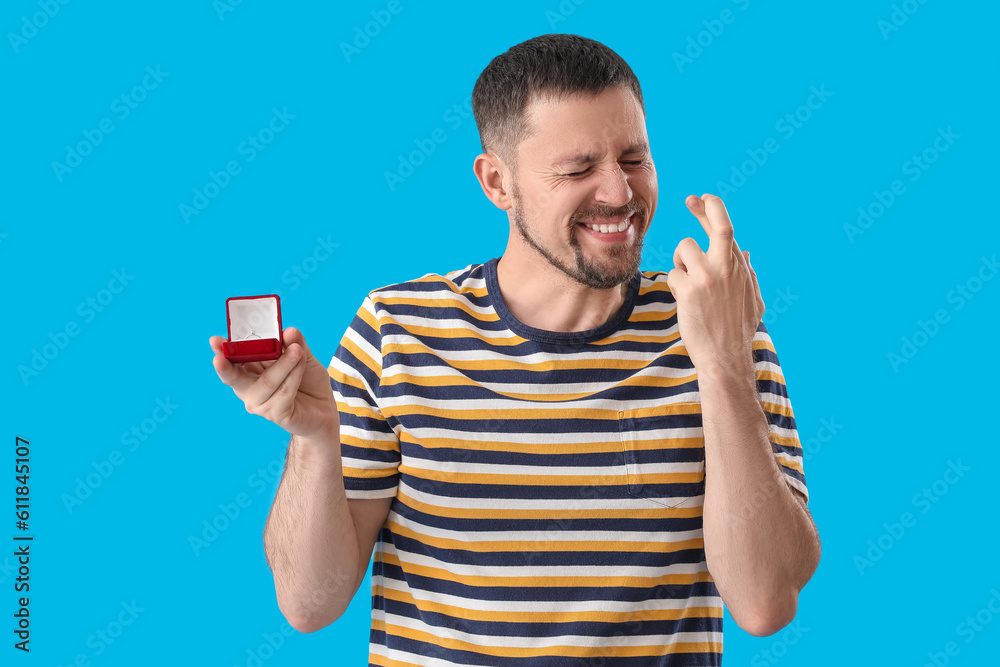 Handsome man with engagement ring crossing fingers on blue background