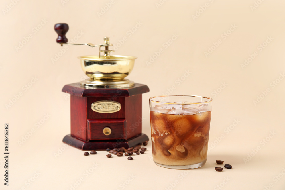 Glass of ice coffee with beans and grinder on beige background