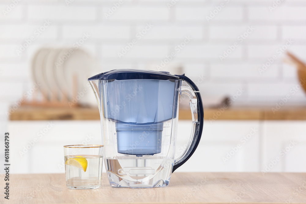 Water filter jug and glass with lemon slice on table in modern kitchen