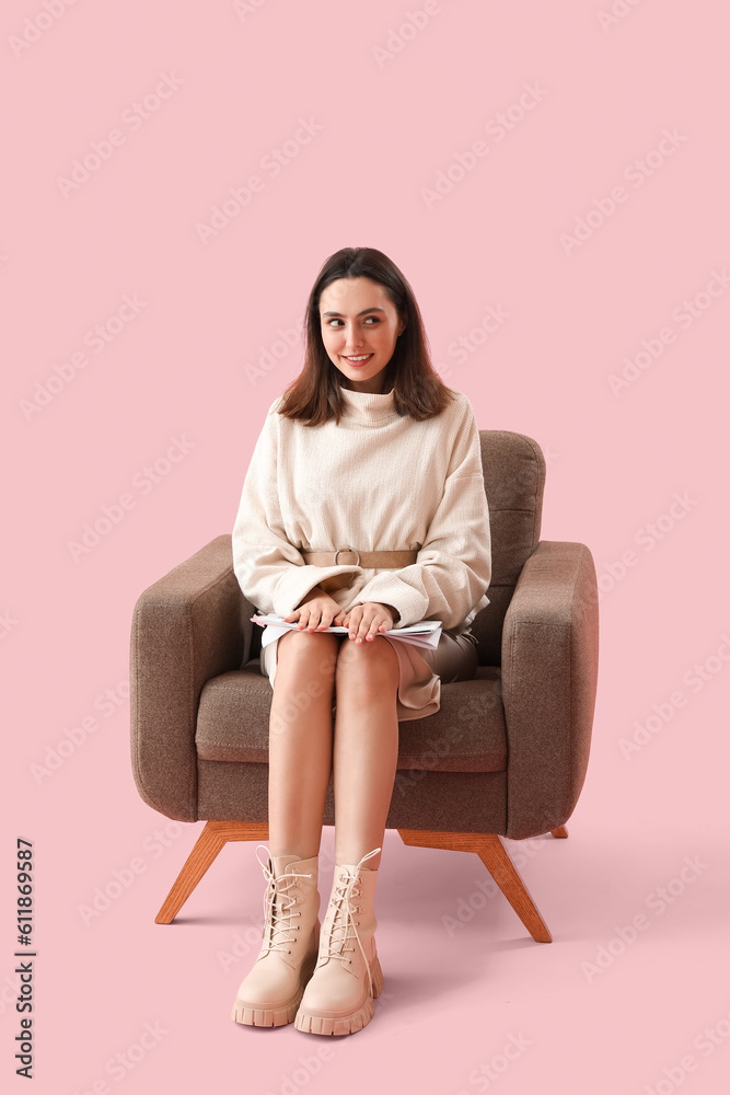 Young woman with newspaper sitting in soft armchair on pink background