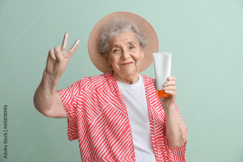 Senior woman with sunscreen cream showing victory gesture on green background