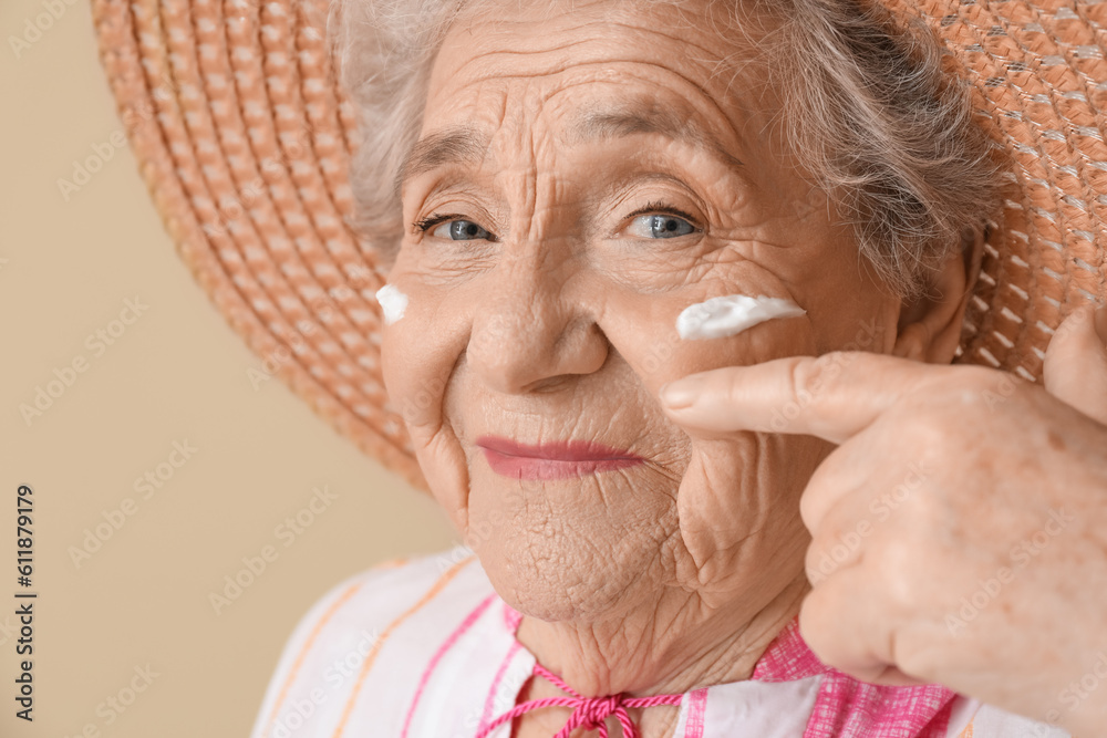 Senior woman with sunscreen cream on her face against beige background, closeup