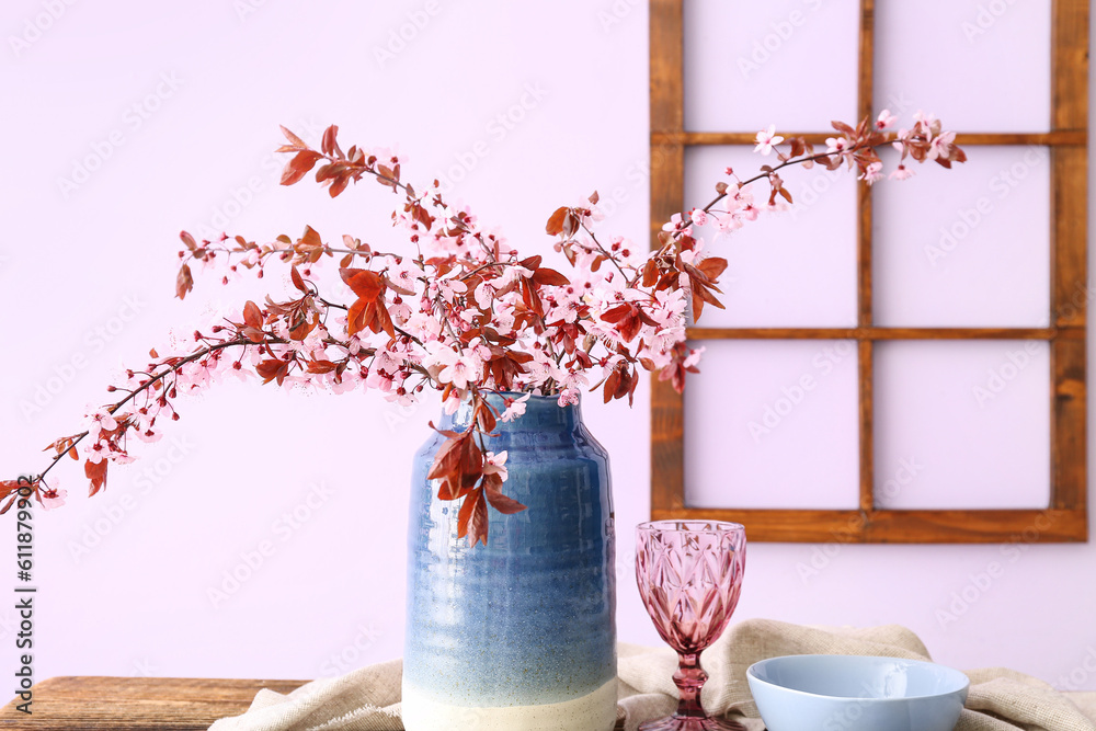 Vase with blooming tree branches, glass, bowl and napkin on wooden table near pink wall