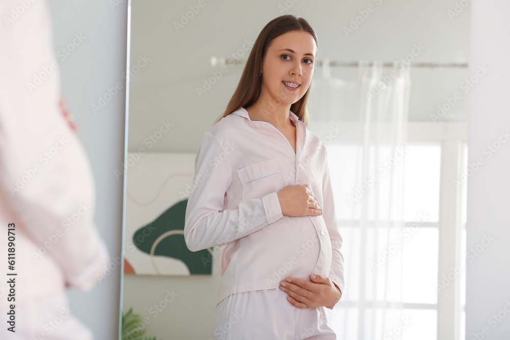 Young pregnant woman in pajamas near mirror at home