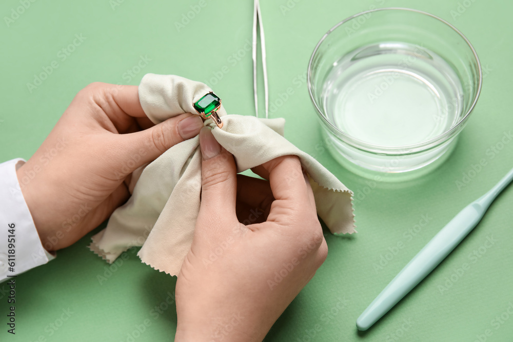 Woman polishing beautiful ring with napkin on green background, closeup