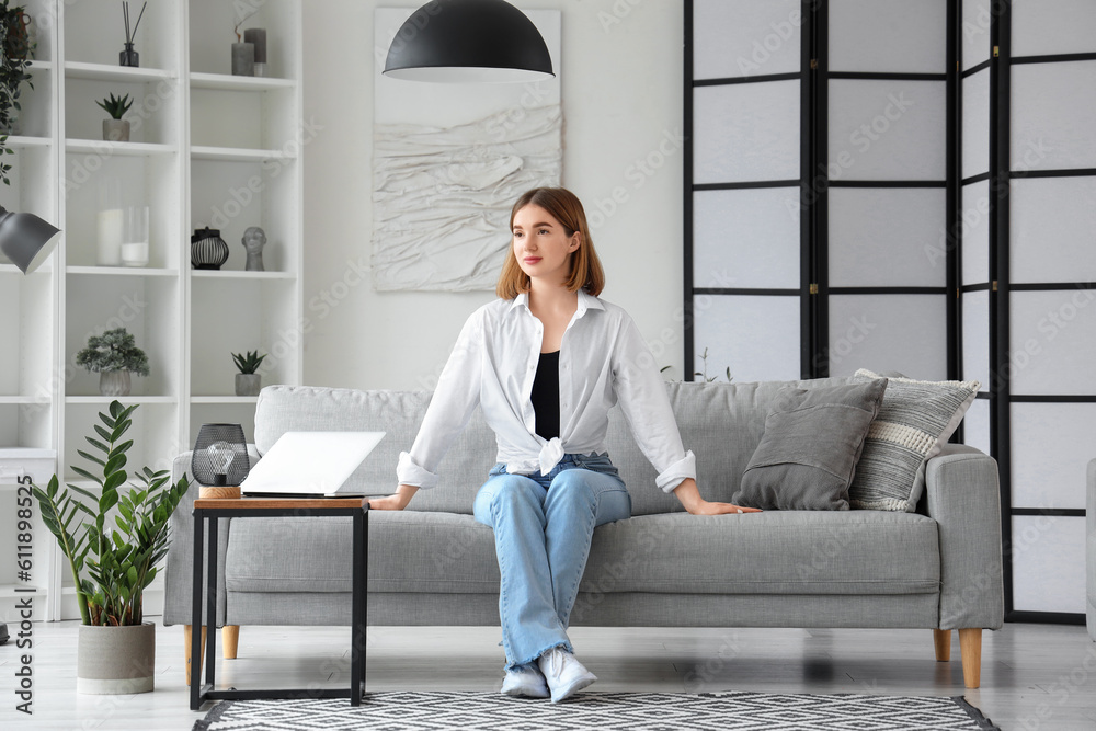 Pretty young woman sitting on grey sofa in interior of light living room