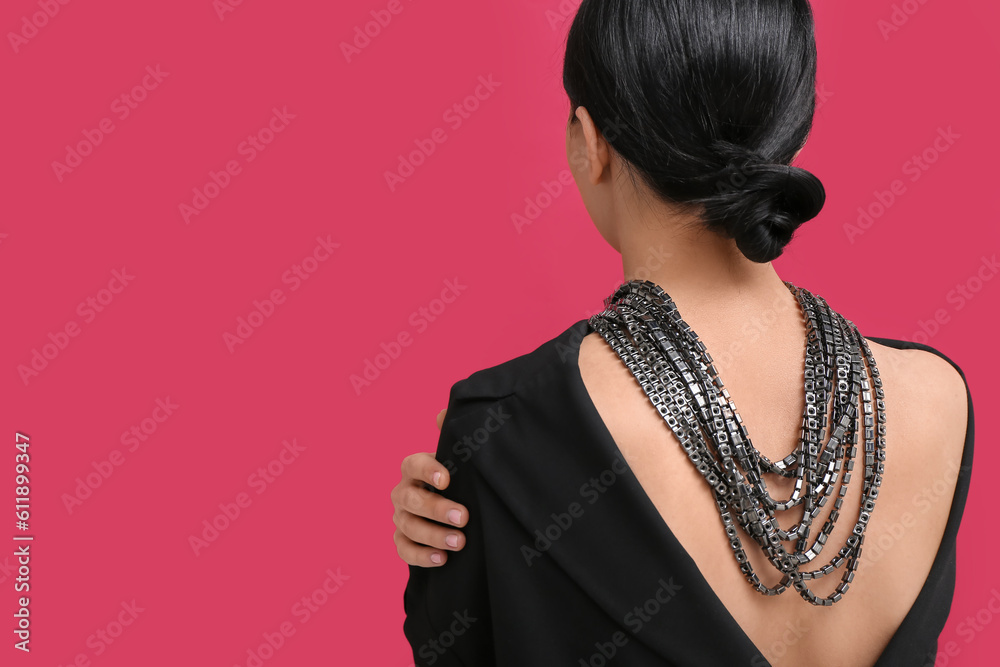 Beautiful young woman wearing massive necklace near red wall, back view