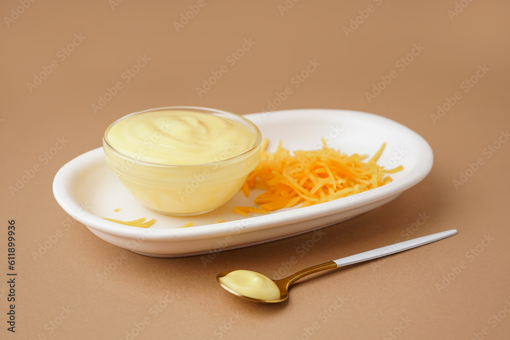 Plate with bowl of tasty cheddar sauce and grated cheese on brown background