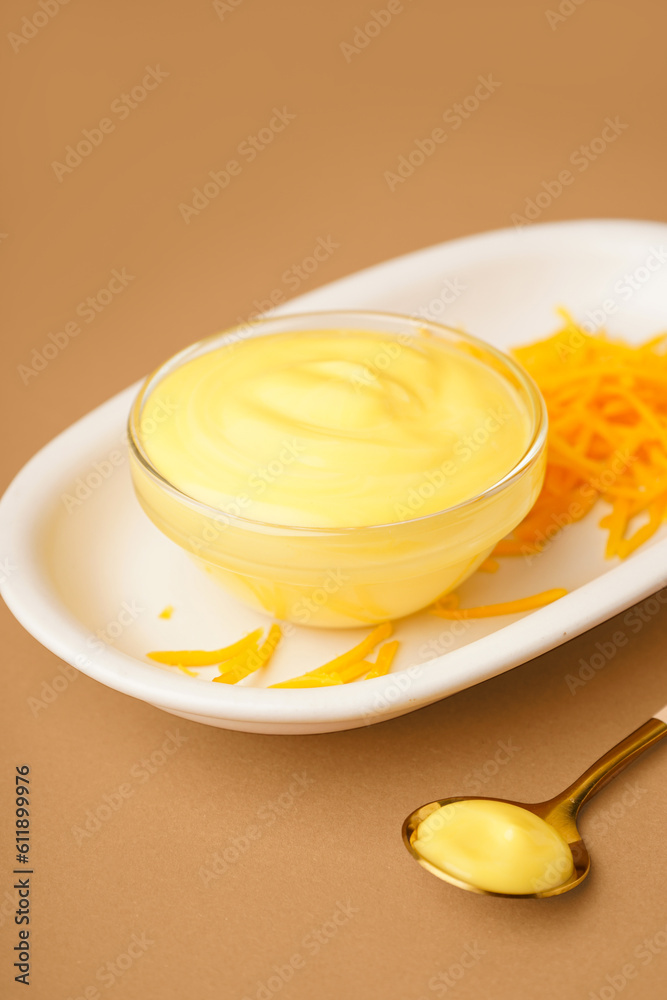 Plate with bowl of tasty cheddar sauce and grated cheese on brown background