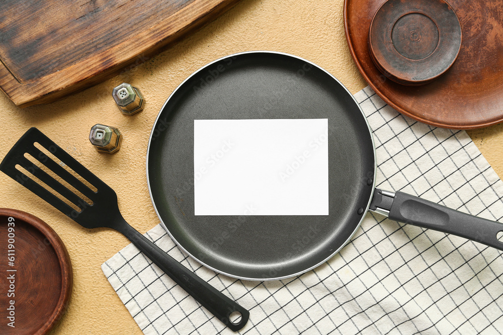 Frying pan with blank card and kitchen utensils on beige background