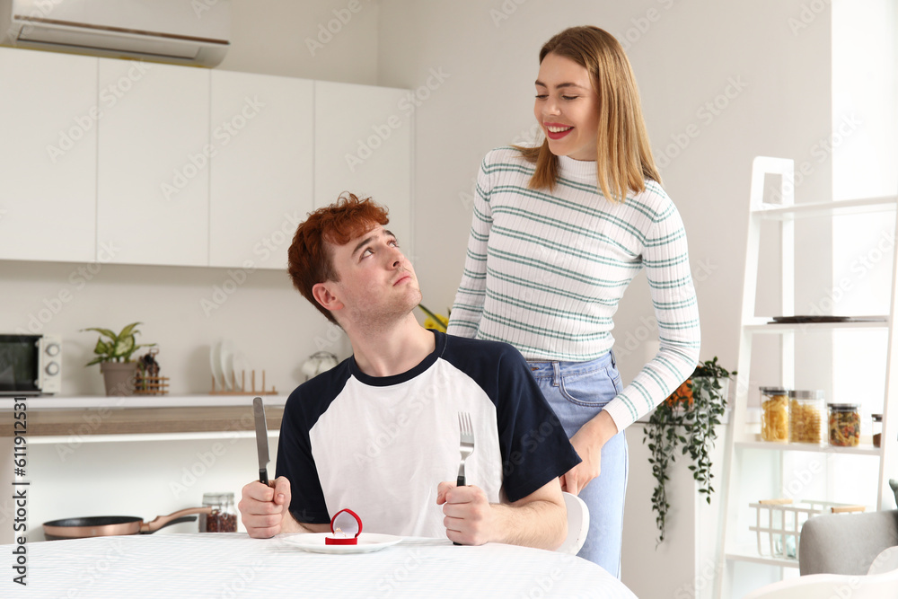 Young woman proposing to her upset boyfriend at table in kitchen