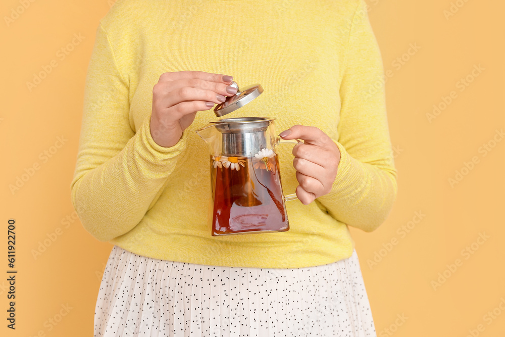 Beautiful young woman with teapot of chamomile tea near yellow wall