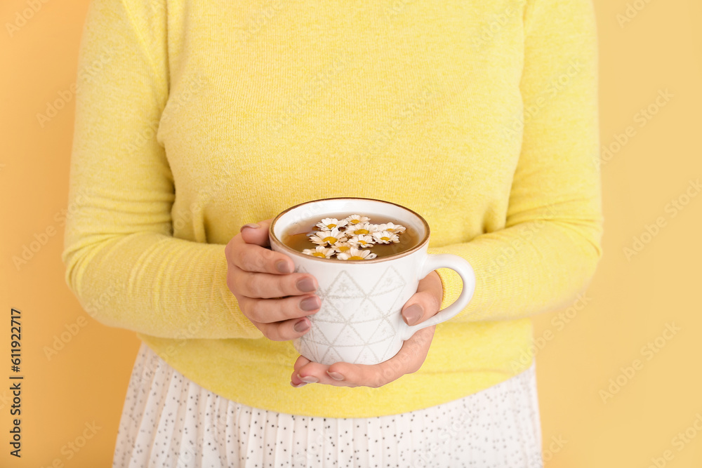 Beautiful young woman with cup of chamomile tea and flowers near yellow wall