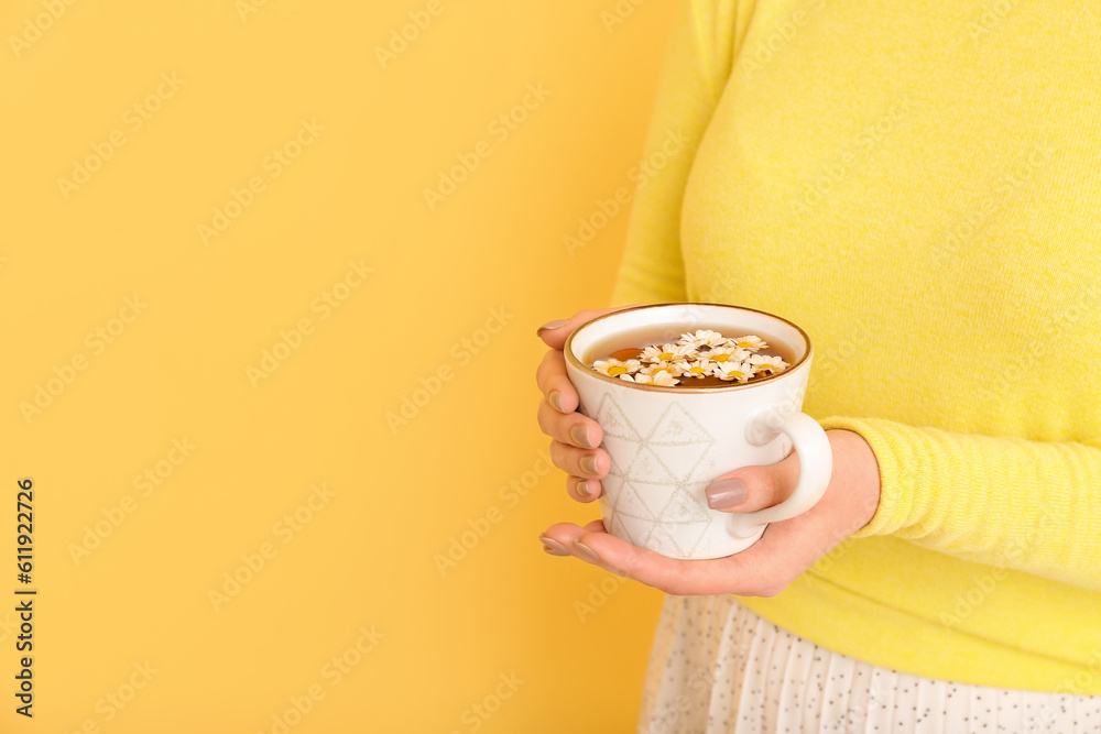 Beautiful young woman with cup of chamomile tea and flowers near yellow wall