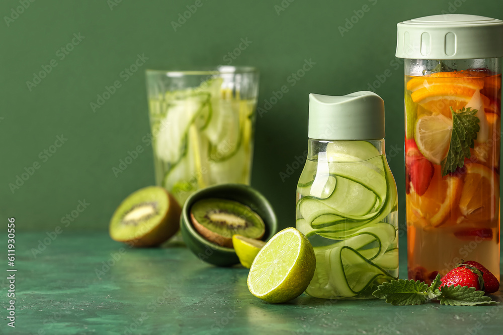 Sports bottles and glass of infused water with different sliced fruits on green background