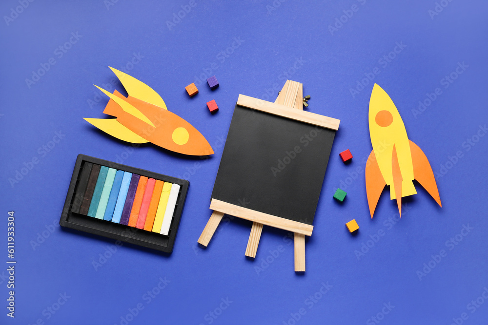 Paper rockets with board, wooden cubes and colorful chalks on blue background