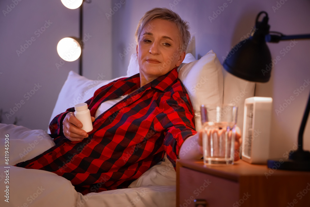 Mature woman with pill bottle taking glass of water from table in bedroom at night