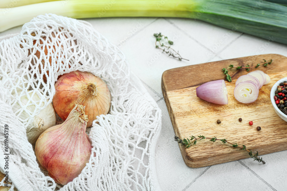 Wooden board and string bag with different kinds of onion on white tile background