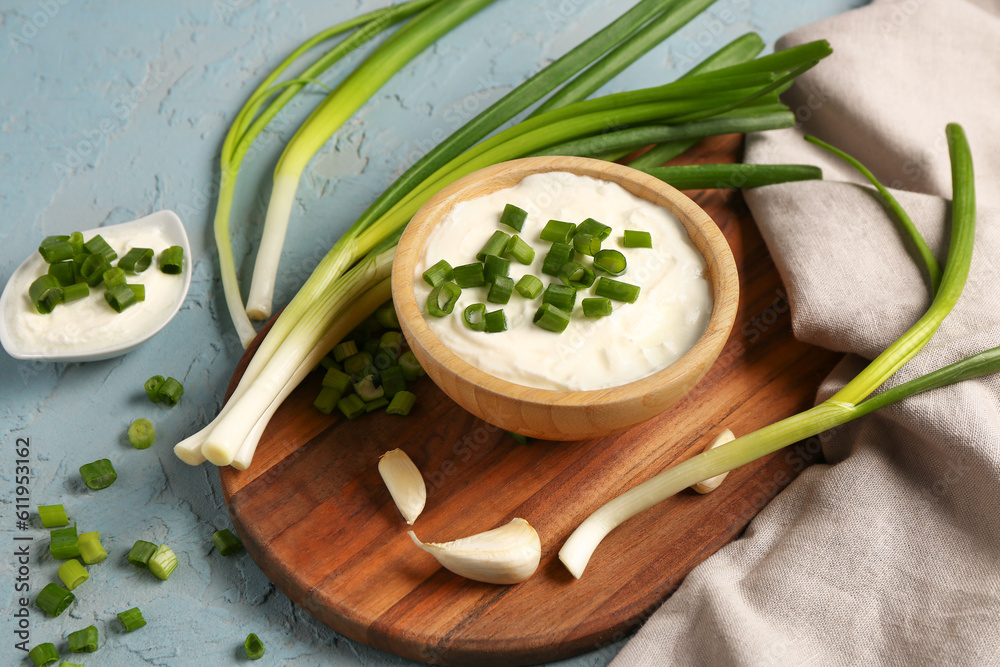 Board and bowl of tasty sour cream with sliced green onion on blue background