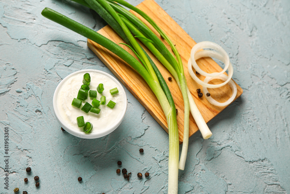 Bowl of tasty sour cream and board with green onion on blue background