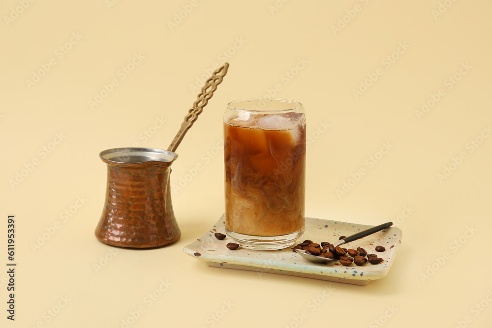 Glass of ice coffee with beans, spoon and pot on yellow background