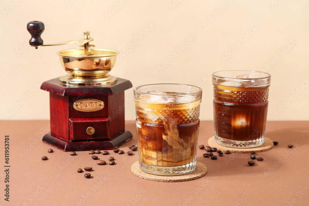 Glasses of ice coffee with beans and grinder on beige table near wall