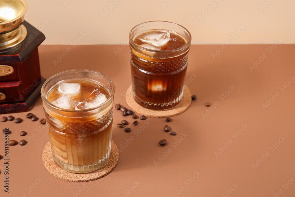 Glasses of ice coffee with beans and grinder on beige table near wall