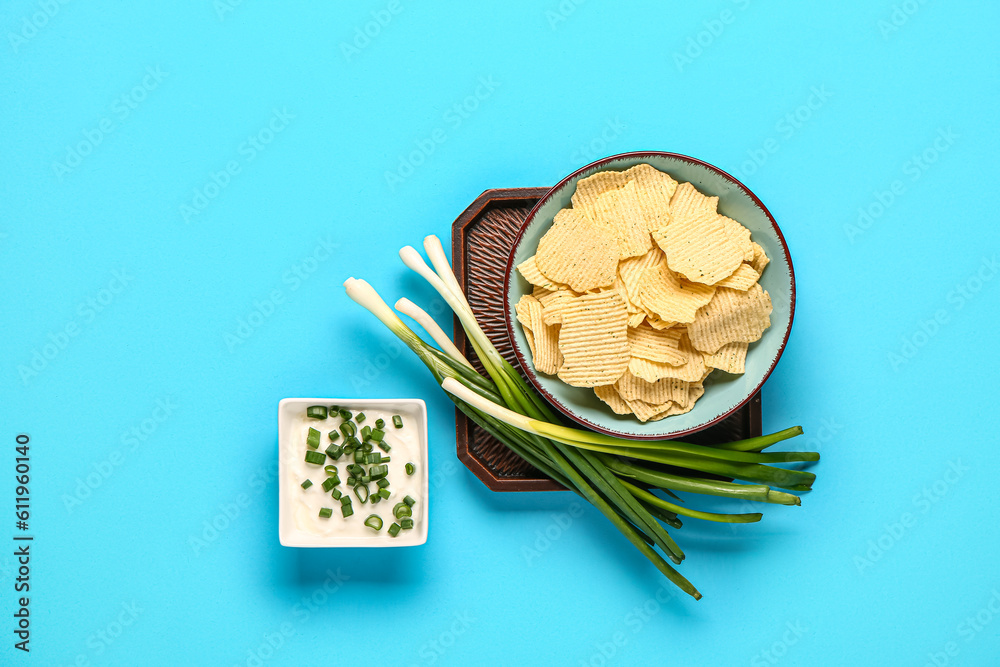 Bowl of tasty sour cream with sliced green onion and potato chips on blue background