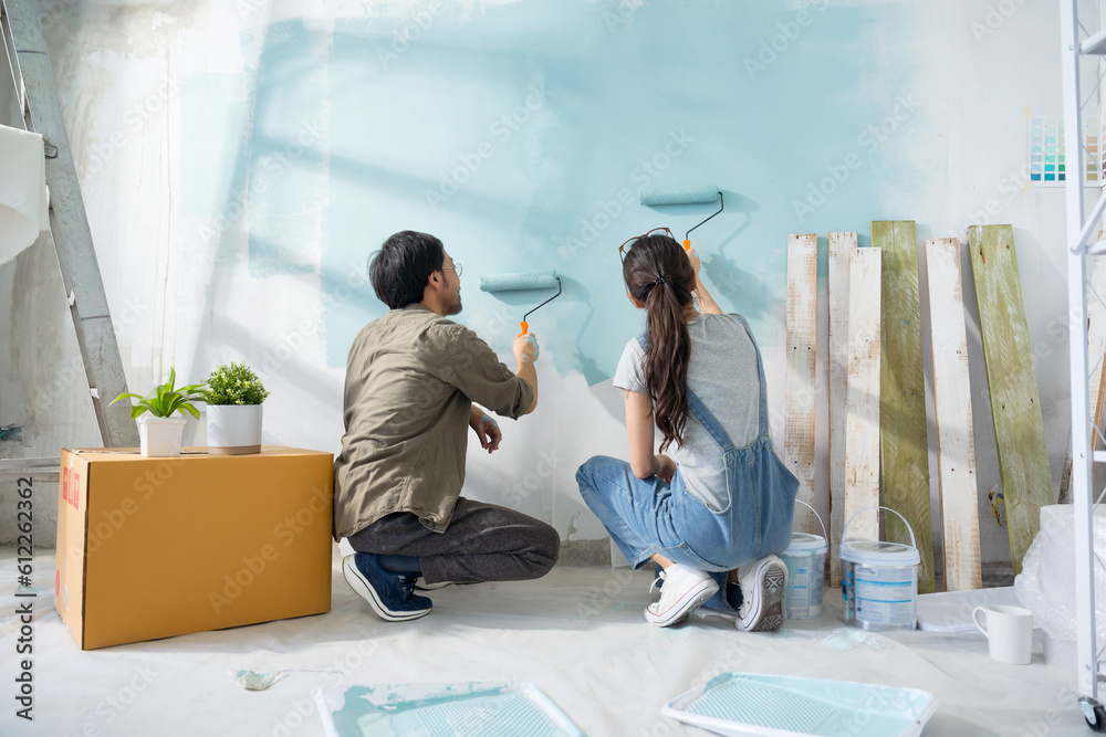 Young Asian couple repairing and painting the wall with blue paint using a roller during renovation 