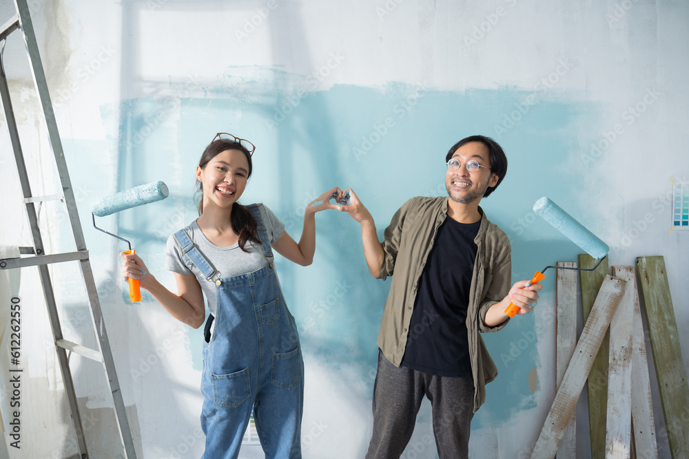 Asian couple expressing love heart while painting the wall with light blue paint using a roller duri