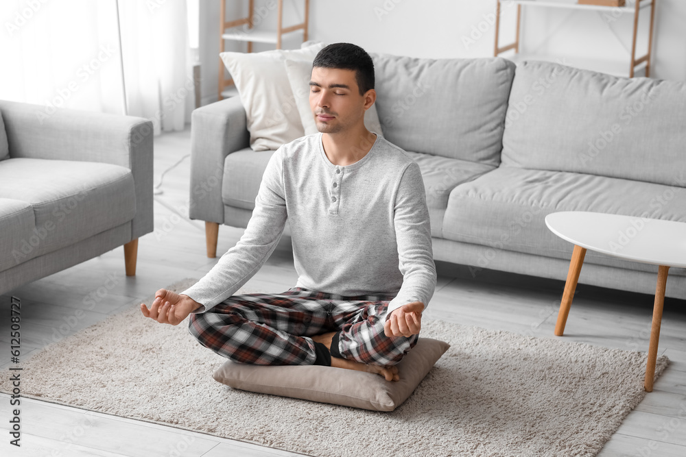 Young man in pajamas meditating on pillow at home