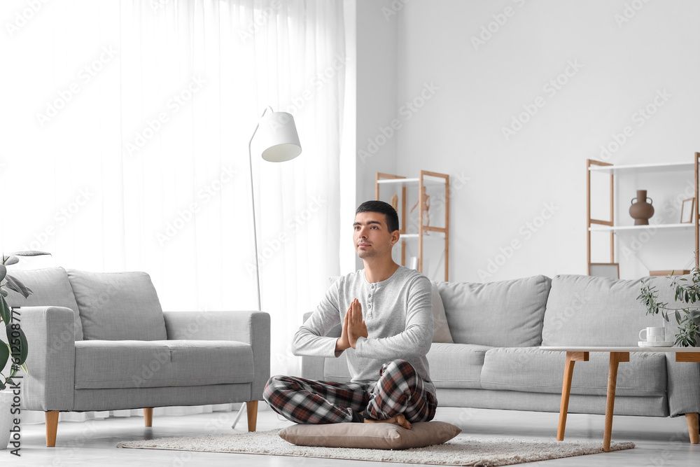 Young man in pajamas meditating on pillow at home