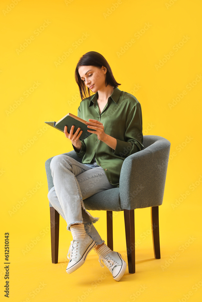 Beautiful woman reading book in soft armchair on yellow background