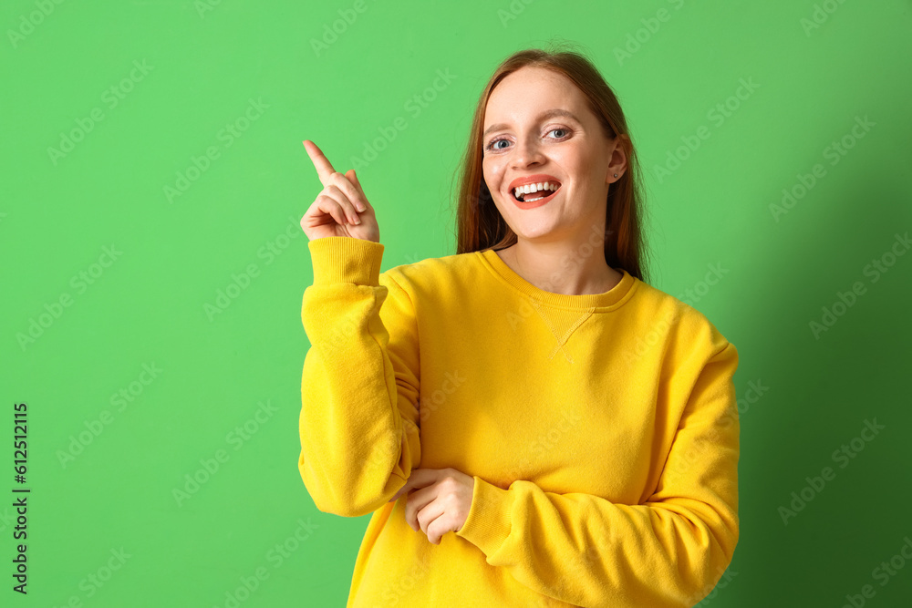 Young redhead woman in sweatshirt pointing at something on green background