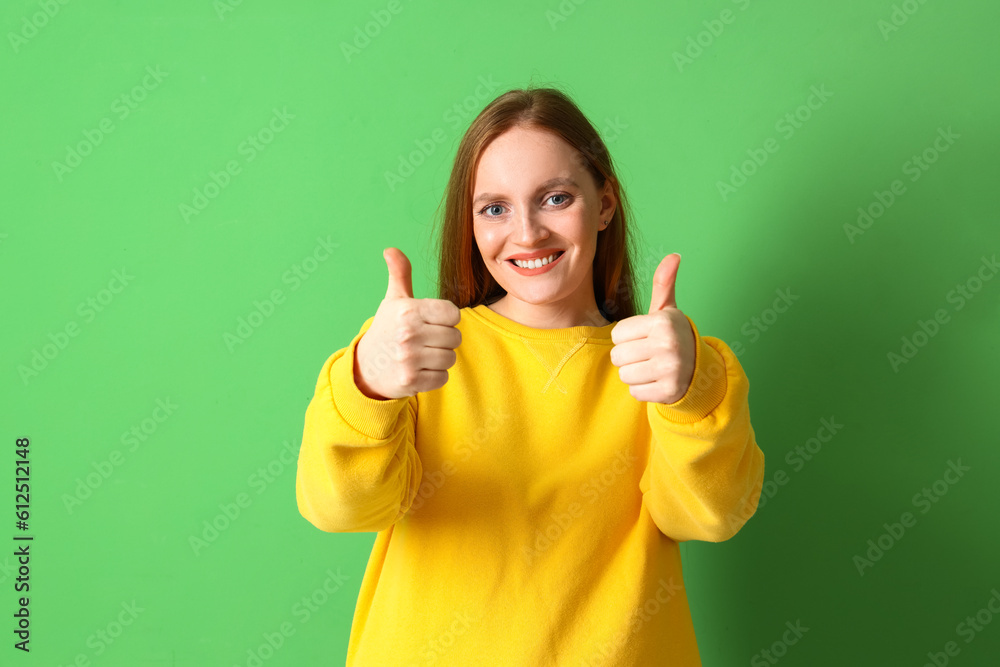 Young redhead woman in yellow sweatshirt showing thumbs-up on green background