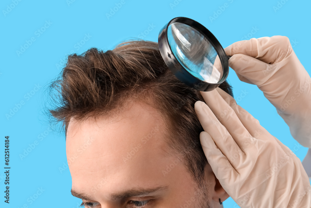 Doctor with magnifier examining young mans hair on blue background, closeup