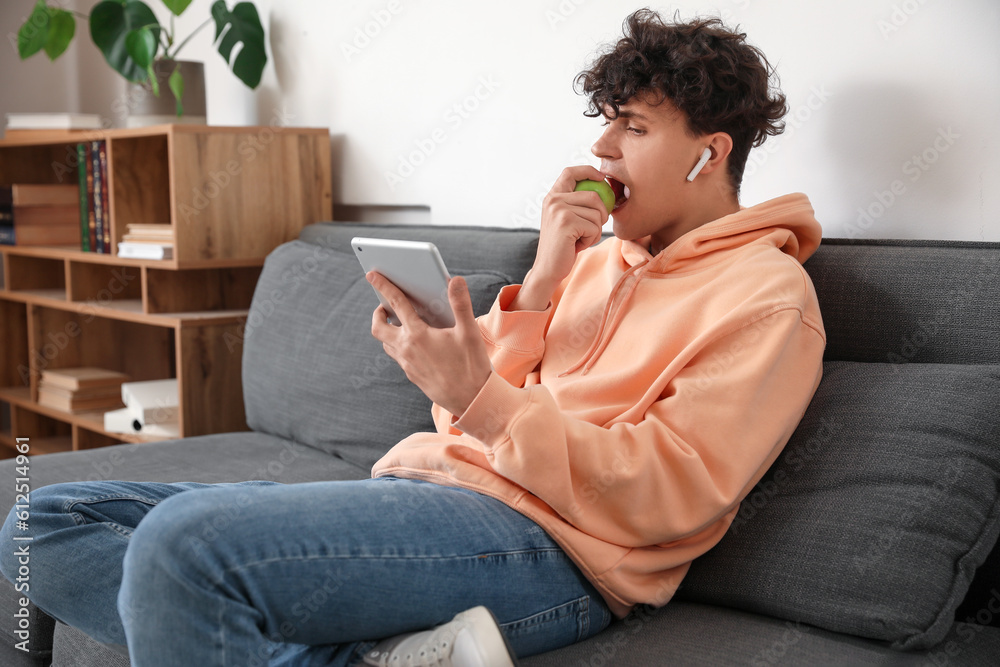 Male student with tablet computer eating apple on sofa at home