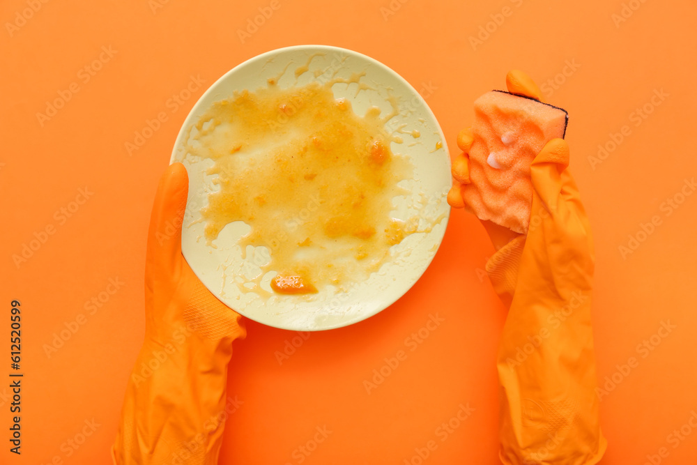 Female hands in rubber gloves washing dirty plate with sponge on orange background